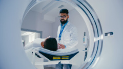 Smiling technician with ID badge standing confidently near MRI tunnel while female patient resting on table. Overseeing post-scan procedure inside professional medical imaging center.